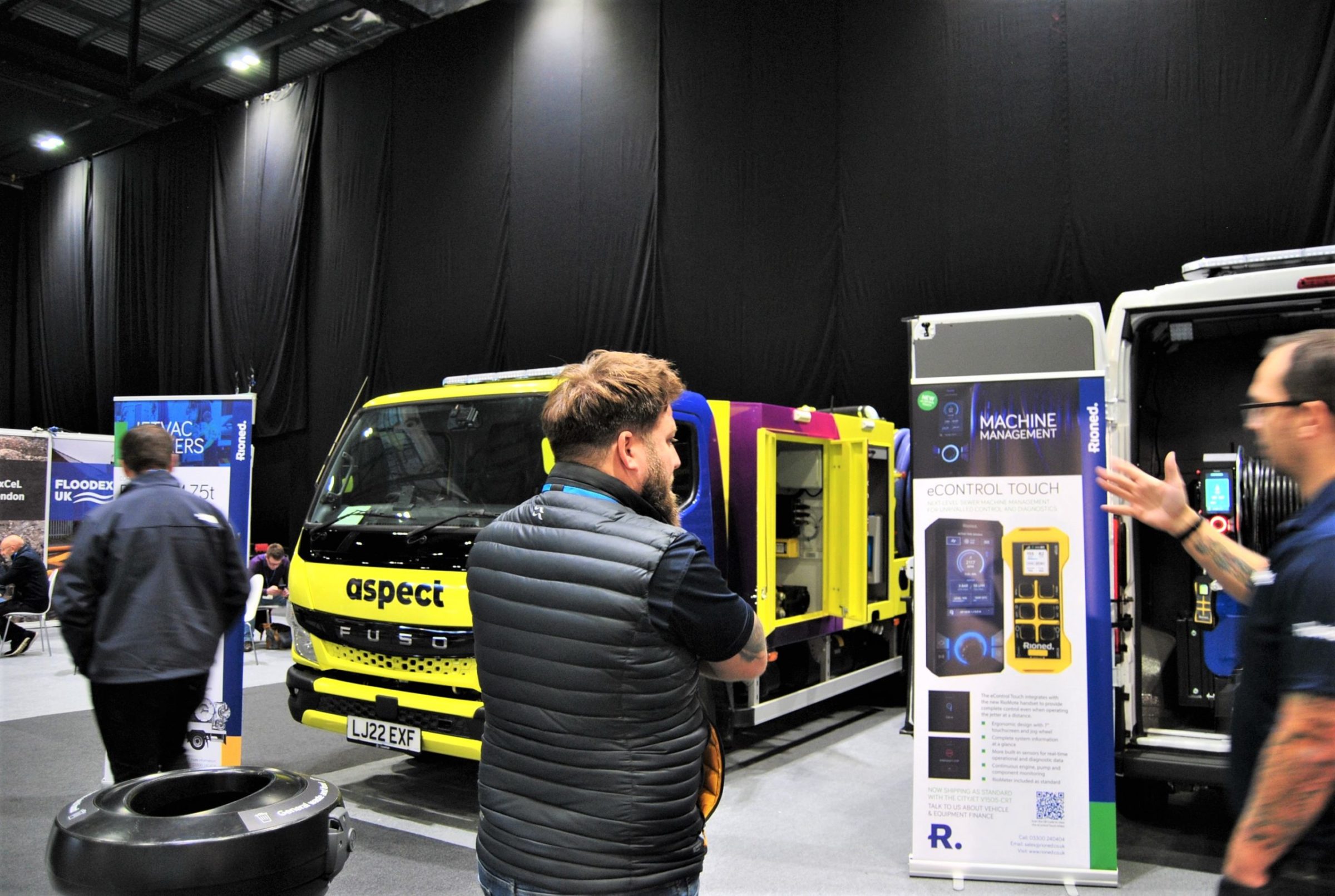 Two men inspect a Rioned van pack jetter, set up in the back of a van. One man is talking to another man who is explaining how it works.